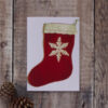Photo of a greetings card on a wooden background. Greetings card shows a red felt Christmas stocking with cream crochet trim.