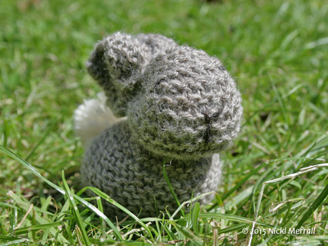 Grey knitted bunny with a cream pompom tail sitting on grass