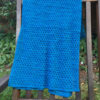 Blue-green rectangular shawl, featuring delicate diamond patterns, draped over wooden chair in garden